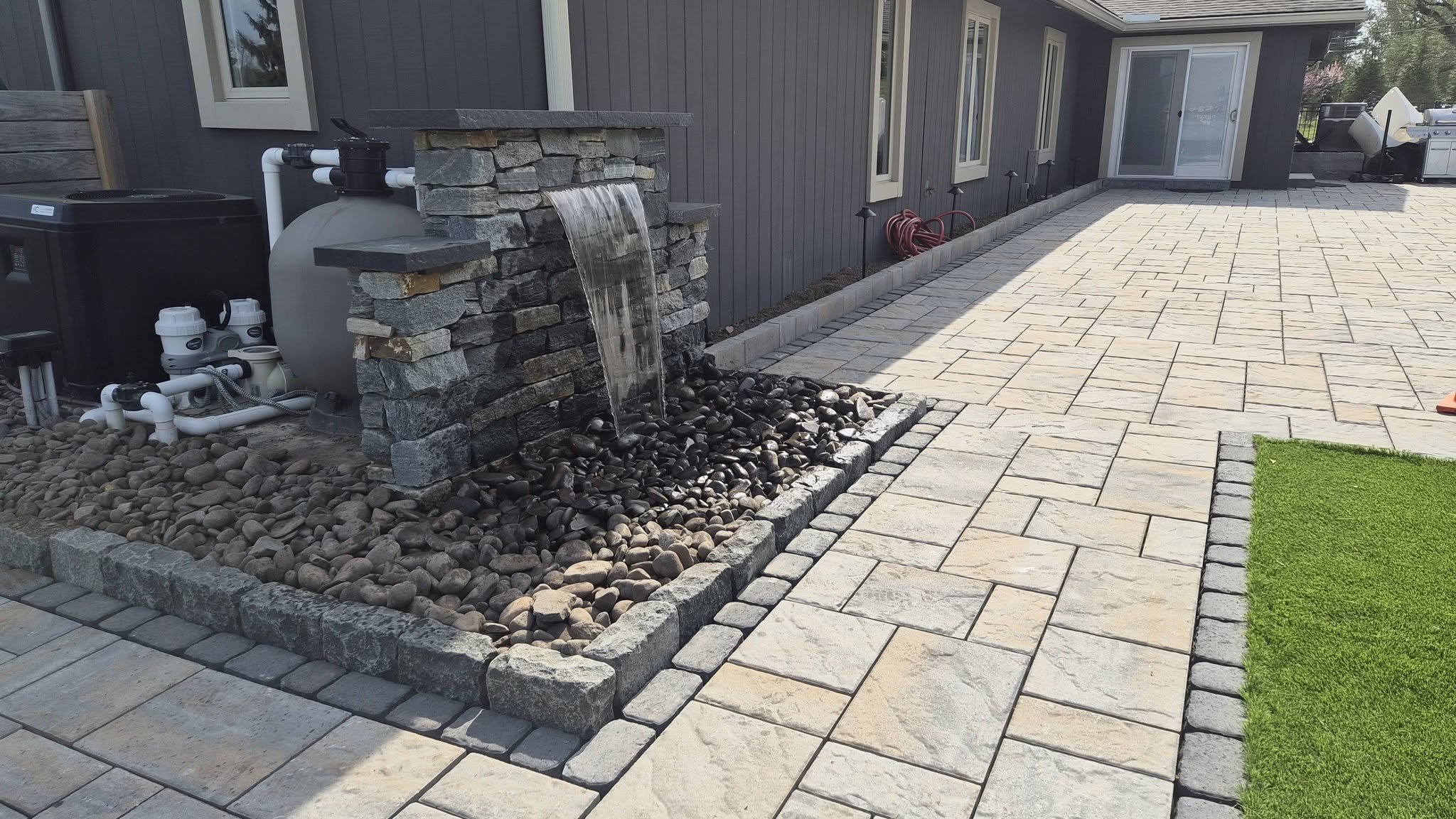 Modern backyard patio featuring a stone paver walkway, integrated stone water feature with spilling waterfall, and clean landscape stone borders next to a lawn. Modern backyard patio featuring a stone paver walkway, integrated stone water feature with spilling waterfall, and clean landscape stone borders next to a lawn.