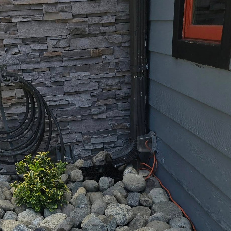 Commercial downspout drainage solution featuring a decorative river rock stone bed next to a gray stone veneer accent wall.